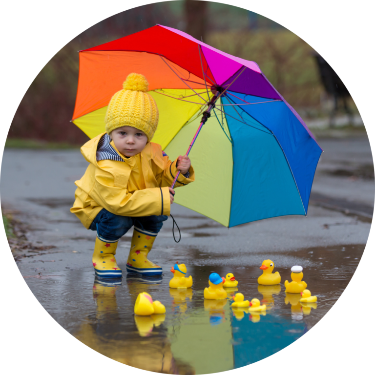 Image d'un enfant accroupi, observant des canards en plastique qui flottent dans une flaque d'eau et tenant un parapluie arc-en-ciel.